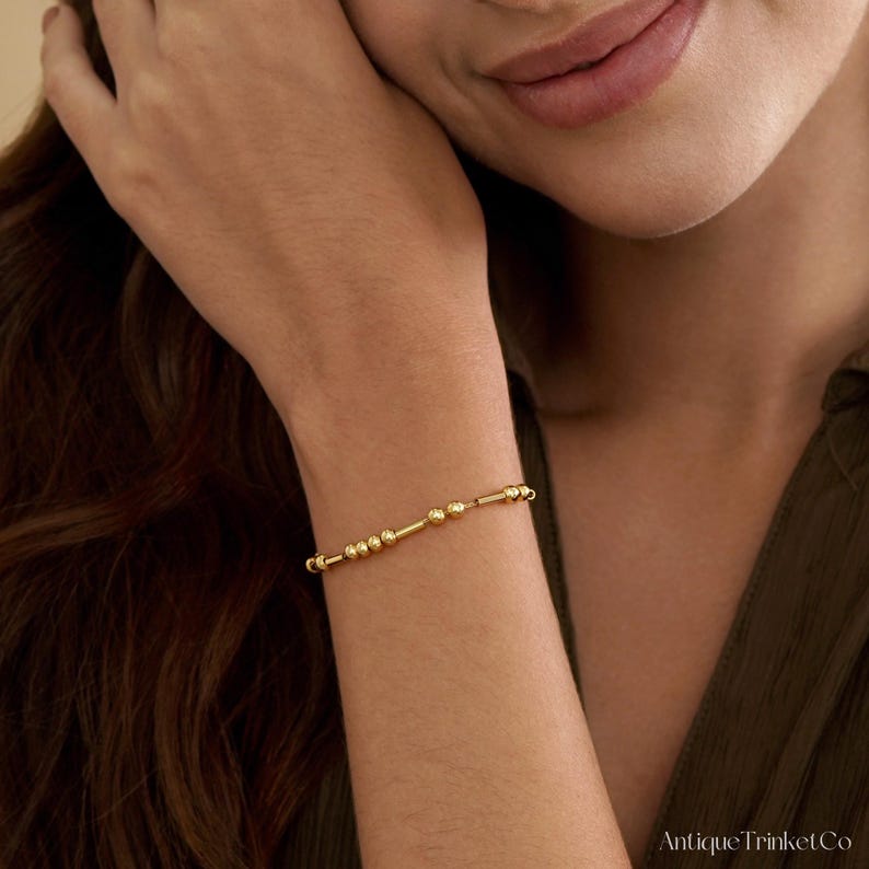 May include: A delicate gold bracelet featuring a series of small, round beads and cylindrical bars. The bracelet is worn on a wrist, showcasing its simple yet elegant design. The bracelet is a piece of jewelry.
