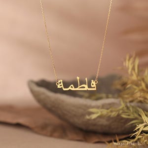 May include: A gold-colored necklace with the name "Fatima" in Arabic script. The pendant hangs from a delicate chain. The background is a soft, blurred mix of beige and brown, with some greenery.