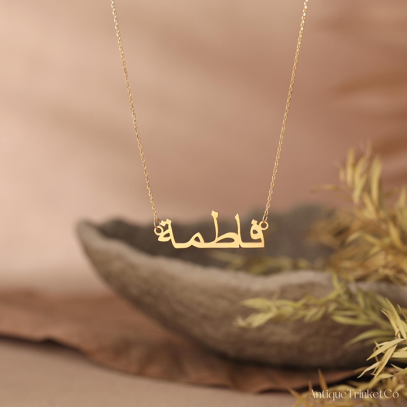 May include: A gold-colored necklace with the name "Fatima" in Arabic script. The pendant hangs from a delicate chain. The background is a soft, blurred mix of beige and brown, with some greenery.