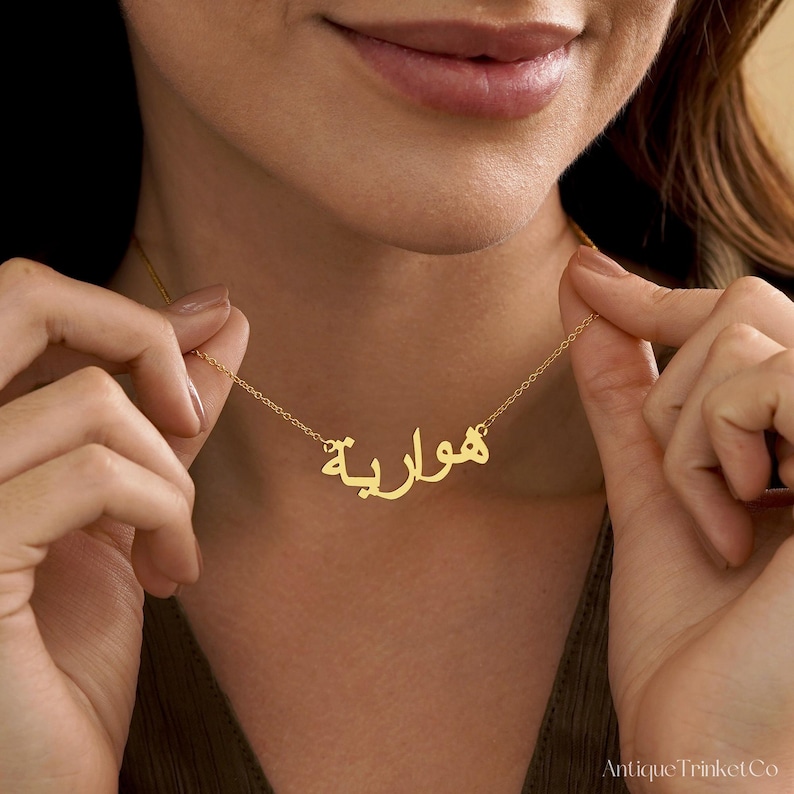 May include: A gold-colored necklace with Arabic script spelling a name. The necklace is held by a person, showcasing the delicate chain and the personalized pendant. The pendant is in a cursive font.