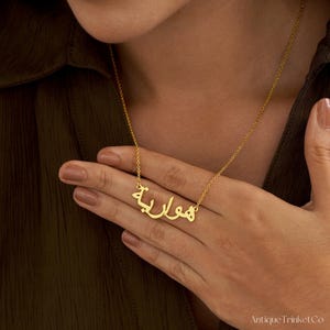 May include: A gold-colored necklace with Arabic script spelling out a name. The necklace is worn around the neck and the person is holding the necklace with their hand. The necklace is on a gold chain.