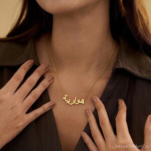 May include: A gold-colored necklace with Arabic script spelling a name. The necklace is worn around the neck, with the chain and pendant visible. The person is wearing a dark green shirt.
