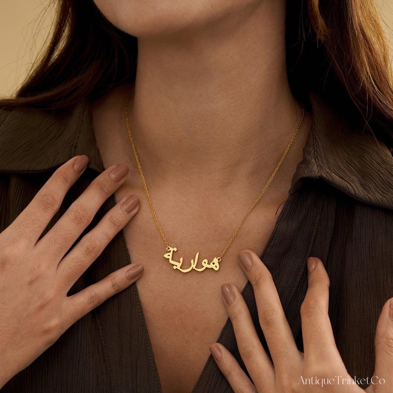 May include: A gold-colored necklace with Arabic script spelling a name. The necklace is worn around the neck, with the chain and pendant visible. The person is wearing a dark green shirt.
