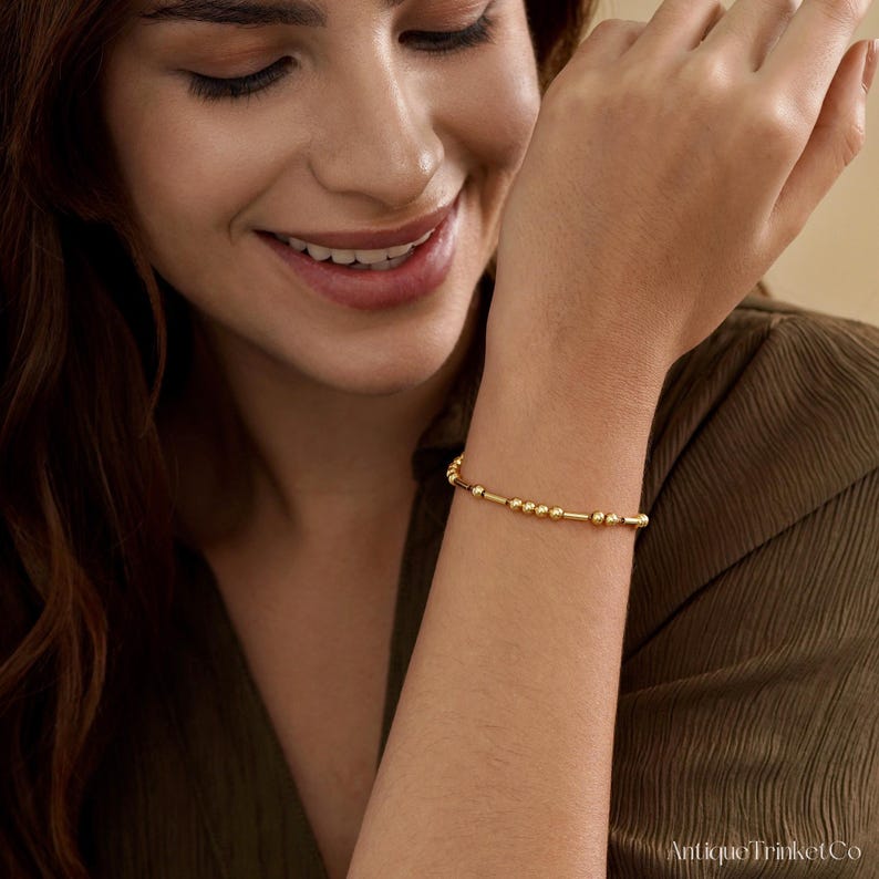 May include: A gold bracelet featuring a series of small, round beads and cylindrical bars. The bracelet is worn on a wrist, showcasing its delicate design and warm, golden hue. The jewelry piece is simple yet elegant.