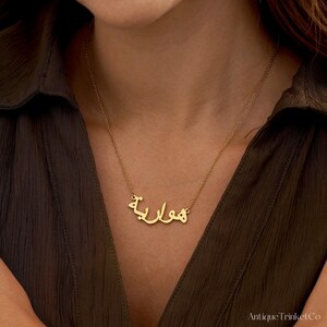 May include: A gold-colored necklace with the Arabic word "هوارية" as a pendant. The necklace is worn around a person's neck, against a dark green shirt. The necklace is delicate and appears to be made of metal.