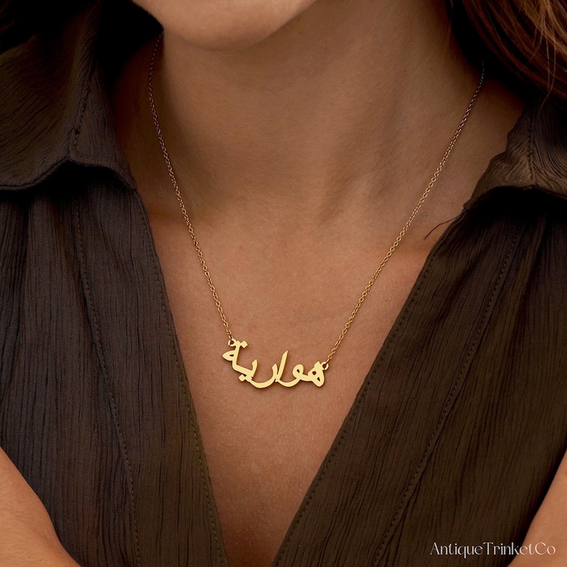 May include: A gold-colored necklace with the Arabic word "هوارية" as a pendant. The necklace is worn around a person's neck, against a dark green shirt. The necklace is delicate and appears to be made of metal.