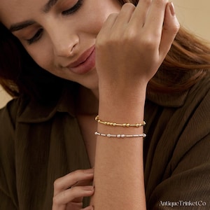 May include: Two delicate bracelets are displayed on a wrist. The top bracelet is gold-toned with small beads and cylindrical bars. The bottom bracelet is silver-toned with a similar design. The bracelets are worn on a person's wrist.