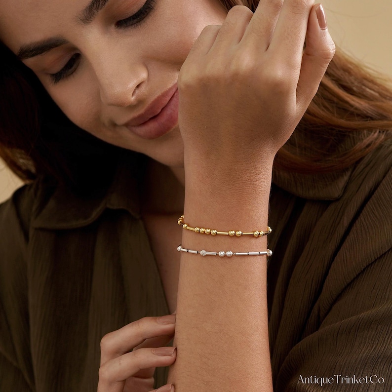 May include: Two delicate bracelets are displayed on a wrist. The top bracelet is gold-toned with small beads and cylindrical bars. The bottom bracelet is silver-toned with a similar design. The bracelets are worn on a person's wrist.