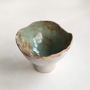 May include: A small, ceramic bowl with a teal glaze and brown accents. The bowl has a unique, irregular shape and a raised base.