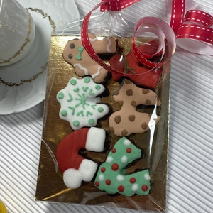 Puede incluir: Un conjunto de seis galletas de jengibre con temática navideña en una bolsa de plástico transparente atada con una cinta roja. Las galletas tienen forma de hombre de jengibre, copo de nieve, árbol de Navidad, calcetín y las letras "J" y "E".
