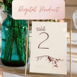May include: A gold-framed table number card with a white background. The card has a brown mountain illustration and the text "table 2" in brown script.