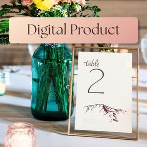Puede incluir: Un marco rectangular de metal dorado que sostiene una tarjeta blanca de número de mesa con la etiqueta 'table 2' y una ilustración de montaña. Un frasco de vidrio verde azulado con flores se sitúa detrás. Una banda rosa dice 'Digital Product'.