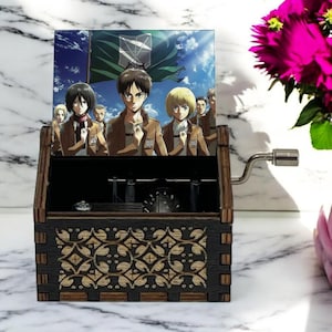May include: Wooden music box with a hand-crank on the side. The lid features a colorful image of characters from the anime Attack on Titan. The characters are standing in front of a green flag with a symbol on it. The music box has a decorative design on the front.
