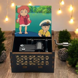 Pode incluir: Uma caixa de m&uacute;sica de madeira com uma manivela no lado. A tampa est&aacute; aberta e apresenta uma ilustra&ccedil;&atilde;o colorida de dois personagens de desenhos animados do filme de anima&ccedil;&atilde;o 'Ponyo'.
