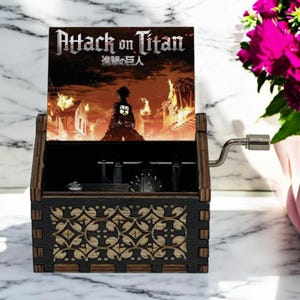May include: A wooden music box with a hand-crank on the side. The lid is open and features a colorful image of a character from the anime Attack on Titan. The text "Attack on Titan" and "進撃の巨人" are printed on the lid.