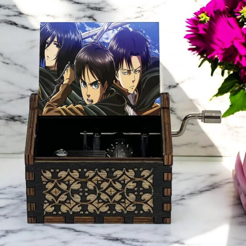 May include: A wooden music box with a hand-crank on the side. The lid of the box features a colorful image of three anime characters from the show Attack on Titan. The box has a decorative design on the front.
