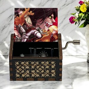 May include: A wooden music box with a hand-crank on the side. The lid is open and features a colorful image of characters from the anime Attack on Titan. The music box has a decorative floral pattern on the front.