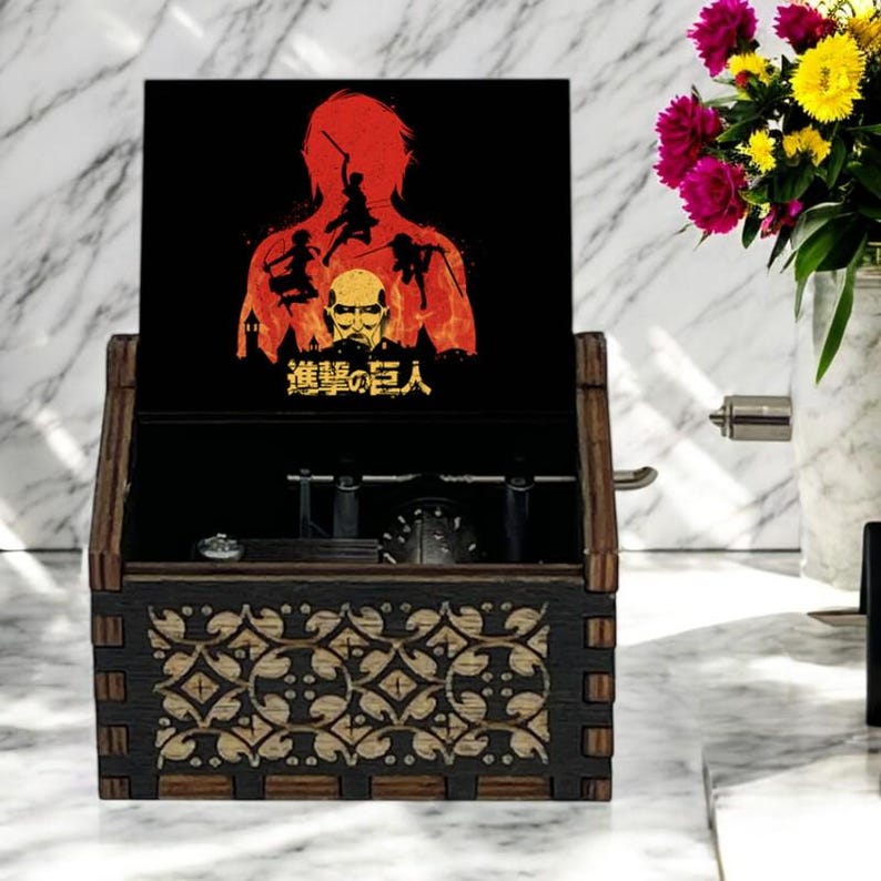 May include: A wooden music box with a black and gold design. The lid is open and features a red and yellow illustration of characters from the anime "Attack on Titan" with the text "進撃の巨人".