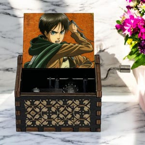 May include: A wooden music box with a hand-crank on the side. The lid features a colorful image of an anime character with brown hair and green eyes, holding a sword. The box has a decorative design on the front.