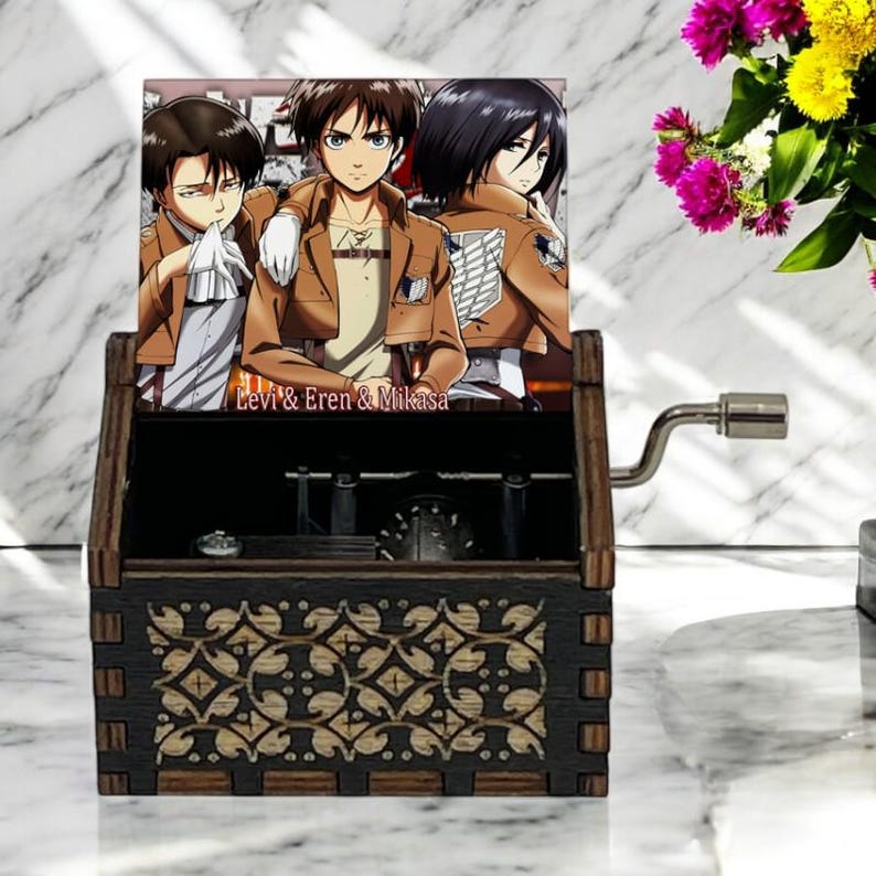 May include: A wooden music box with a hand-crank on the side. The top of the box features a colorful image of three anime characters from the show Attack on Titan. The image is captioned "Levi & Eren & Mikasa".