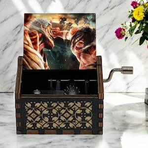 May include: A wooden music box with a hand-crank on the side. The lid of the box features a colorful image of two characters from the anime Attack on Titan. The box has a decorative floral pattern on the front.