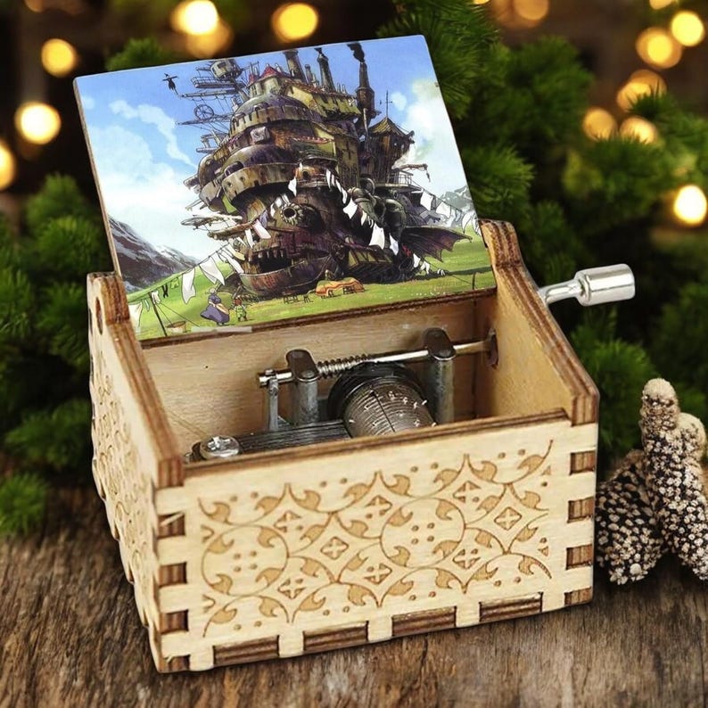 May include: A wooden music box with a hand-crank mechanism. The lid is open and features a printed image of a flying airship from the animated film 'Howl's Moving Castle'. The box has a decorative carved pattern on the front.