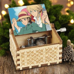 May include: A wooden music box with a hand-crank on the side. The lid is open and features a printed image of two animated characters, a woman with brown hair and a man with blonde hair, standing together. The music box has a decorative carved design on the front.