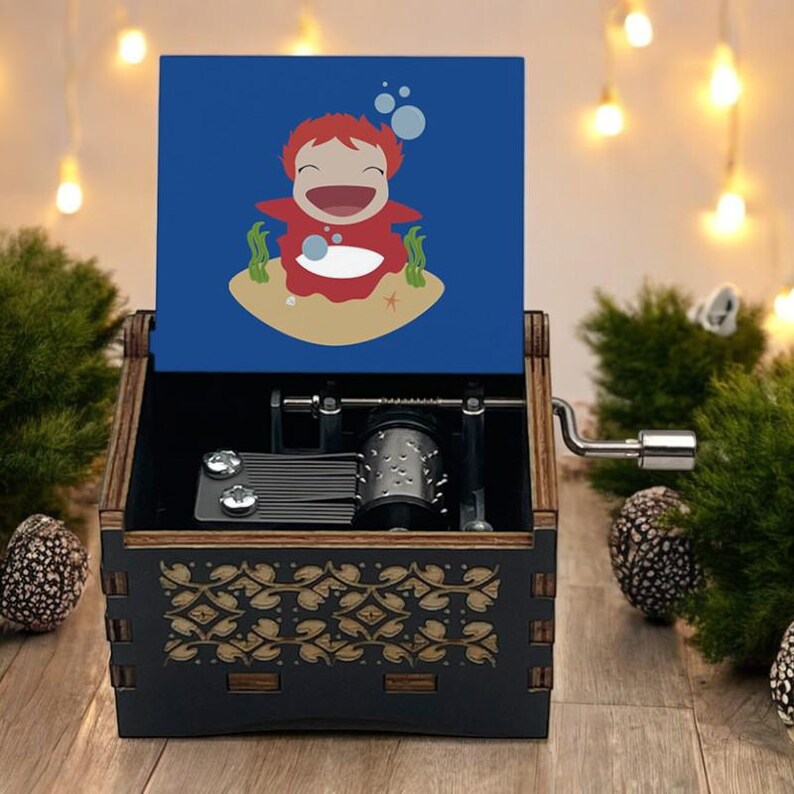 Pode incluir: Uma caixa de m&uacute;sica de madeira com uma tampa azul que apresenta um personagem de desenho animado com cabelo vermelho e um vestido vermelho. O personagem est&aacute; sorrindo e soprando bolhas. A caixa de m&uacute;sica tem uma manivela no lado.