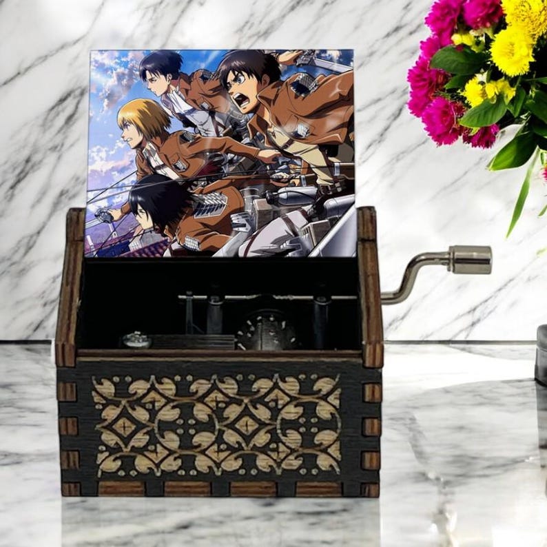 May include: A wooden music box with a hand-crank on the side. The lid is open and features a colorful image of characters from the anime Attack on Titan. The music box has a decorative design on the front.