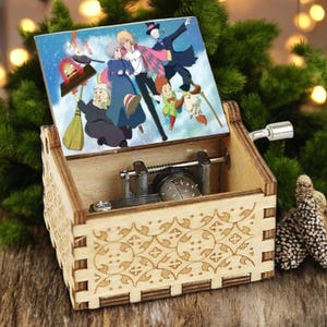 May include: A wooden music box with a hand-crank on the side. The lid is open and features a colorful illustration of characters from the animated film "Howl's Moving Castle".