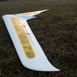Può includere: Un'ala di aeromodello bianca e gialla è appoggiata su un campo erboso. L'ala ha un design curvo con una sezione gialla e diversi piccoli elementi di fissaggio neri. L'ala è angolata, suggerendo che potrebbe essere stata usata di recente.