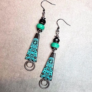 May include: Teal and black dangle earrings with a geometric pattern and two silver rings. The earrings have a black bead and a teal bead.
