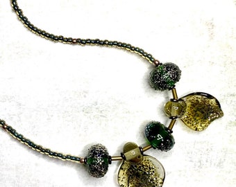 May include: A green and gold beaded necklace with a clasp fastening. The necklace features several large, irregularly shaped green and gold glass beads.
