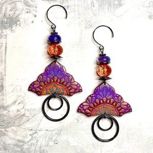 May include: A pair of dangle earrings with a purple, orange, and gray mandala design. The earrings have a black wire loop and a hook closure. The beads are orange, purple, and gold.