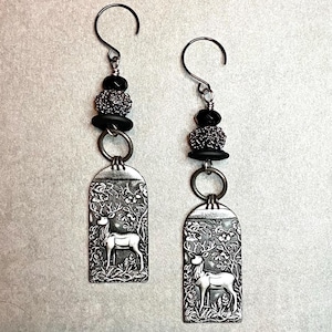 May include: A pair of silver dangle earrings with black beads and a silver charm of a deer in a forest scene.