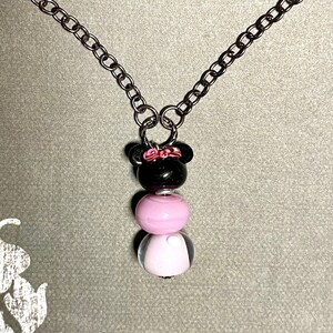 May include: A silver chain necklace with a black and pink bead charm. The charm is shaped like a cartoon character with a bow on its head.