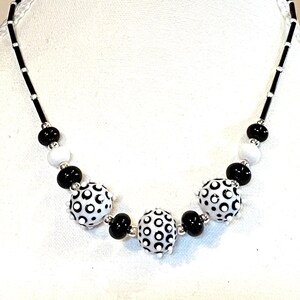 May include: A black and white necklace with large, round beads. The beads are white with a black polka dot pattern. The necklace is on a white mannequin.