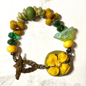 Puede incluir: Una pulsera artesanal con una variedad de cuentas en tonos verdes, amarillos y dorados. La pulsera incluye una cuenta en forma de p&aacute;jaro y un cierre en forma de flor. La pulsera mide aproximadamente 17.78 cm de largo.
