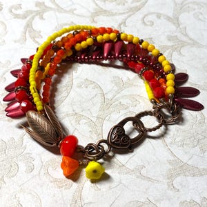 Op de afbeelding: Een armband met meerdere strengen, voorzien van gele, oranje en rode kralen, geaccentueerd met donkerrode, bloembladachtige kralen. Het sieraad bevat een koperkleurige, bladvormige connector, een hartvormige karabijnsluiting en kleine bloemvormige bedels.