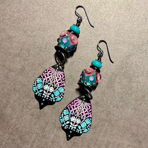 May include: A pair of dangle earrings with black metal teardrop-shaped charms featuring a white and blue floral design. The charms are attached to a beaded section with pink, turquoise, and purple beads.