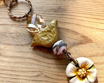 May include: A gold bird charm with a white and black bead and a white flower charm with a yellow center hangs from a brown chain with clear glass beads.