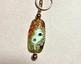 May include: A gold-toned keychain with a glass bead featuring a detailed illustration of a woman's face with orange and brown swirls. The bead is attached to a gold-toned chain and a split ring.
