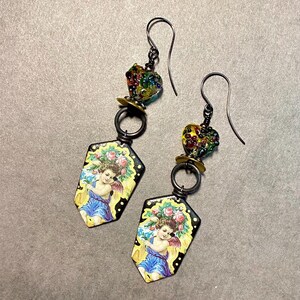 May include: A pair of dangle earrings featuring a vintage-style design. Each earring has a black metal hook, a colorful glass bead, and a pendant with an image of a cherub. The pendants are yellow with a black border and a floral design.