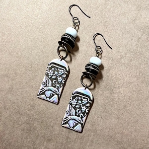 May include: A pair of dangle earrings with a silver-toned metal charm featuring a floral design and a bird. The charms are attached to a black bead and a white bead. The earrings are made with a silver-toned metal hook.