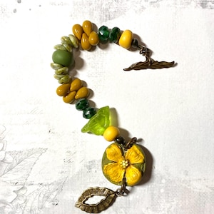 Puede incluir: Una pulsera hecha a mano con cuentas amarillas, verdes y oliva. La pulsera incluye cuentas en forma de l&aacute;grima, cuentas redondas y un dije en forma de flor. El cierre es un cierre de palanca de color bronce.