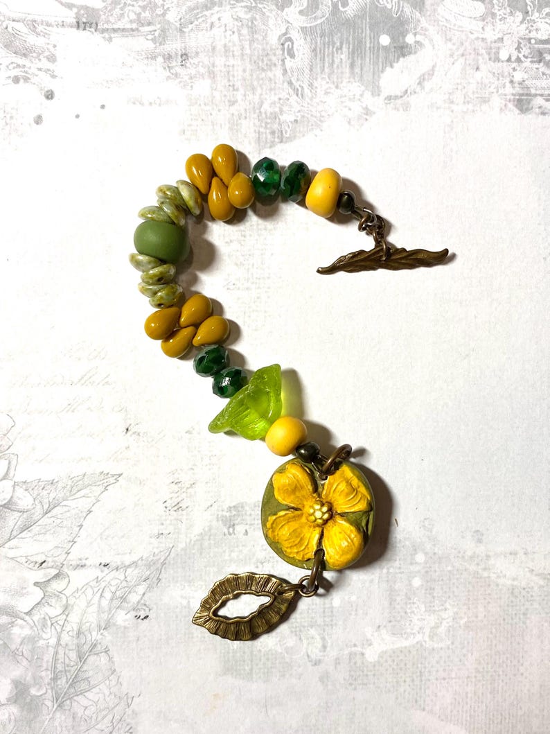 Puede incluir: Una pulsera hecha a mano con cuentas amarillas, verdes y oliva. La pulsera incluye cuentas en forma de l&aacute;grima, cuentas redondas y un dije en forma de flor. El cierre es un cierre de palanca de color bronce.
