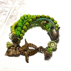 Könnte beinhalten: Ein mehrreihiges Armband mit grünen und blauen Glasperlen, bronzefarbenen Blattanhängern und Blumenakzenten.