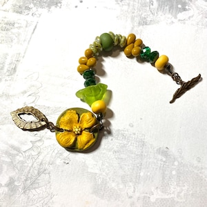 Puede incluir: Una pulsera hecha a mano con un dise&ntilde;o floral. La pulsera est&aacute; compuesta por varias cuentas en tonos amarillos, verdes y transparentes. Una gran flor amarilla es el punto focal, conectada a un cierre con dise&ntilde;o de hoja.
