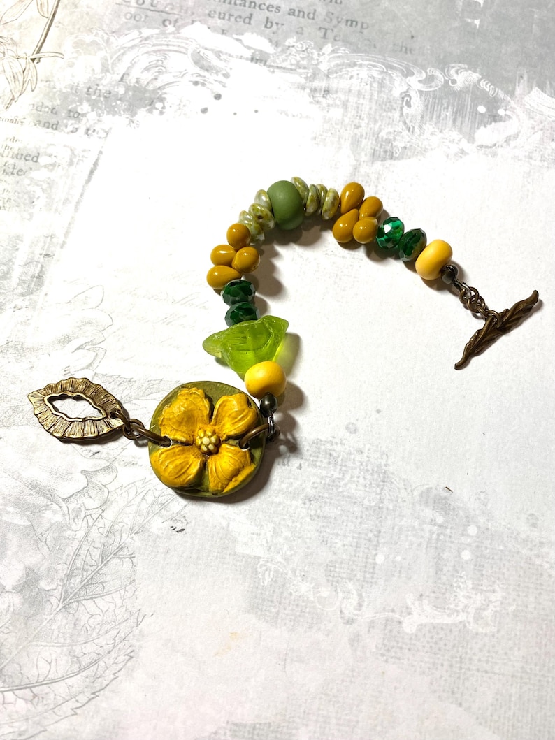 Puede incluir: Una pulsera hecha a mano con un dise&ntilde;o floral. La pulsera est&aacute; compuesta por varias cuentas en tonos amarillos, verdes y transparentes. Una gran flor amarilla es el punto focal, conectada a un cierre con dise&ntilde;o de hoja.