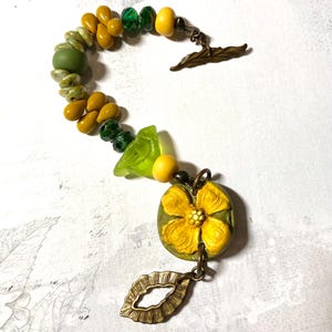 Puede incluir: Un brazalete artesanal con una mezcla de cuentas amarillas, verdes y transparentes. El brazalete incluye un dije de flor amarilla detallada y un cierre en forma de hoja de bronce. Las cuentas son de varias formas y tama&ntilde;os.
